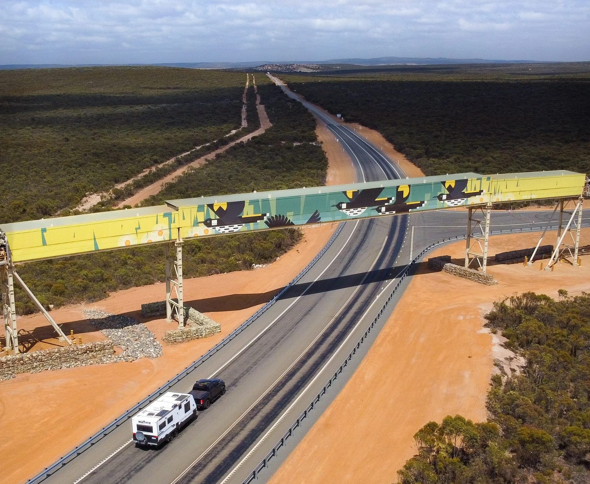 An aerial view of a highway passing under an enclosed overpass painted with a bright geometric mural of birds, with a camper van driving below.
