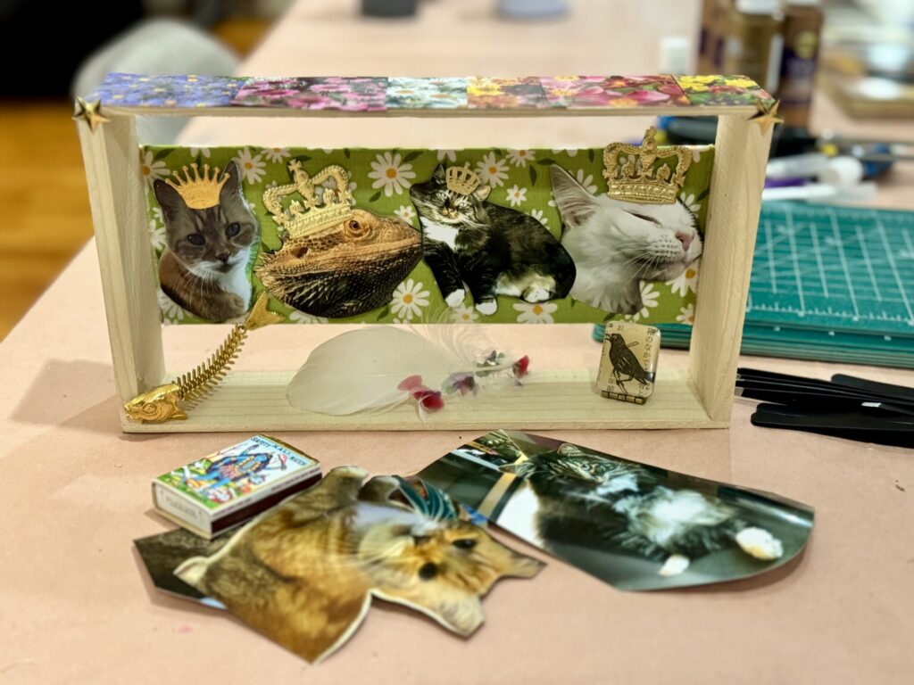A wooden shadow box holding cutout photos of three cats and a bearded dragon, each wearing a small gold crown, against green daisy fabric.