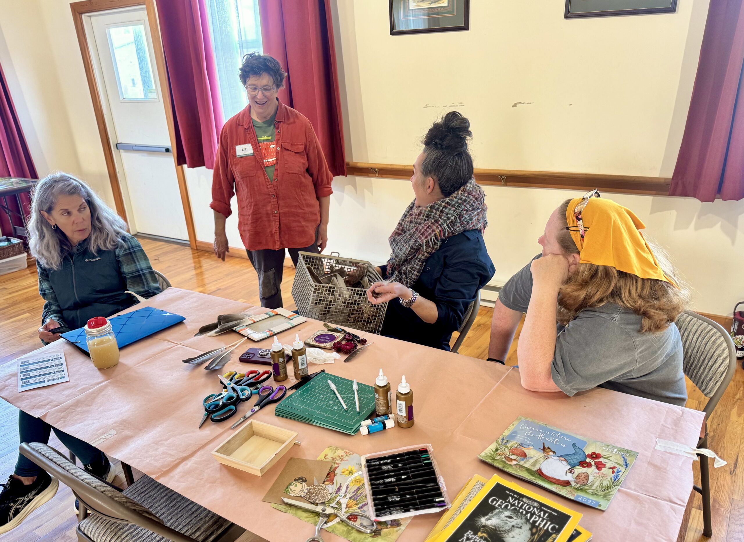 Liz Fox, standing and laughing, talks with three seated participants at a table of scissors, feathers, glue, markers, and magazines.