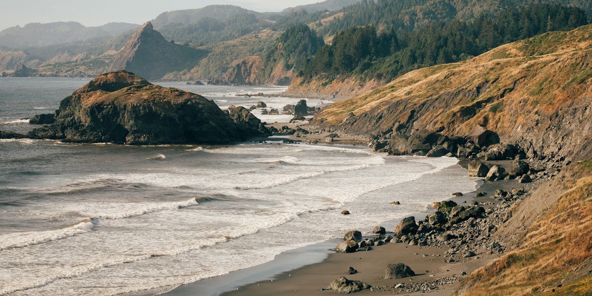 Surf rolling onto a rocky Oregon Coast beach below forested headlands and a sea stack.